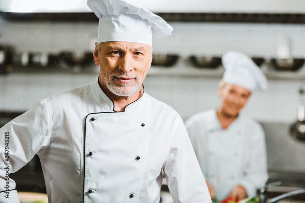handsome male chef in uniform and cap looking at camera in restaurant kitchen with copy space