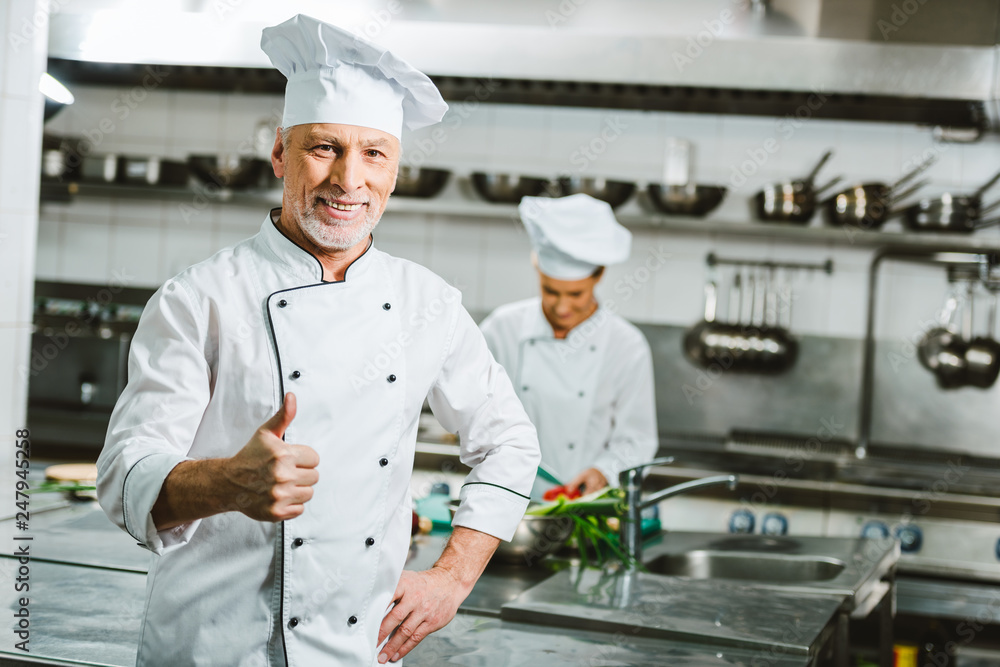 handsome smiling male chef in uniform looking at camera and showing thumb up sign in restaurant kitchen