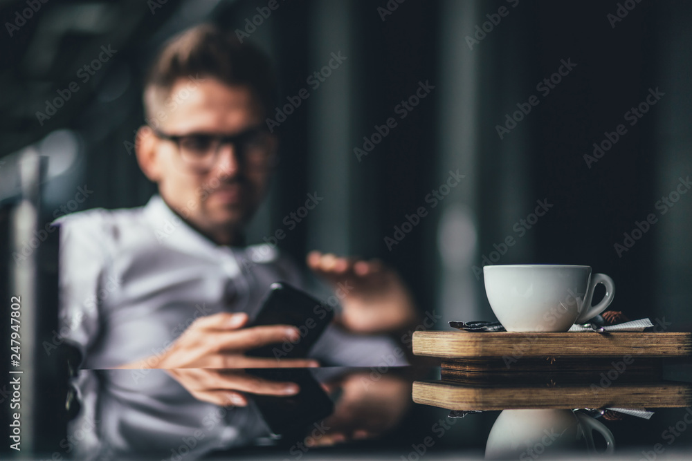 Young businessman using phone while cup of coffee stand on the table in cafe. Focus on cup Young businessman using phone while cup of coffee stand on the table in cafe. Focus on cup