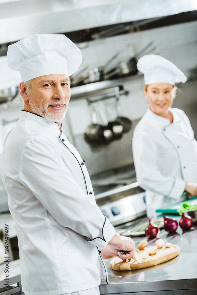 handsome male chef looking at camera while cooking with female colleague in restaurant kitchen