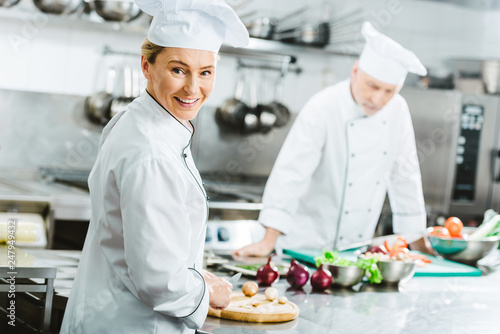 beautiful female chef looking at camera and smiling while cooking with colleague in restaurant kitchen
