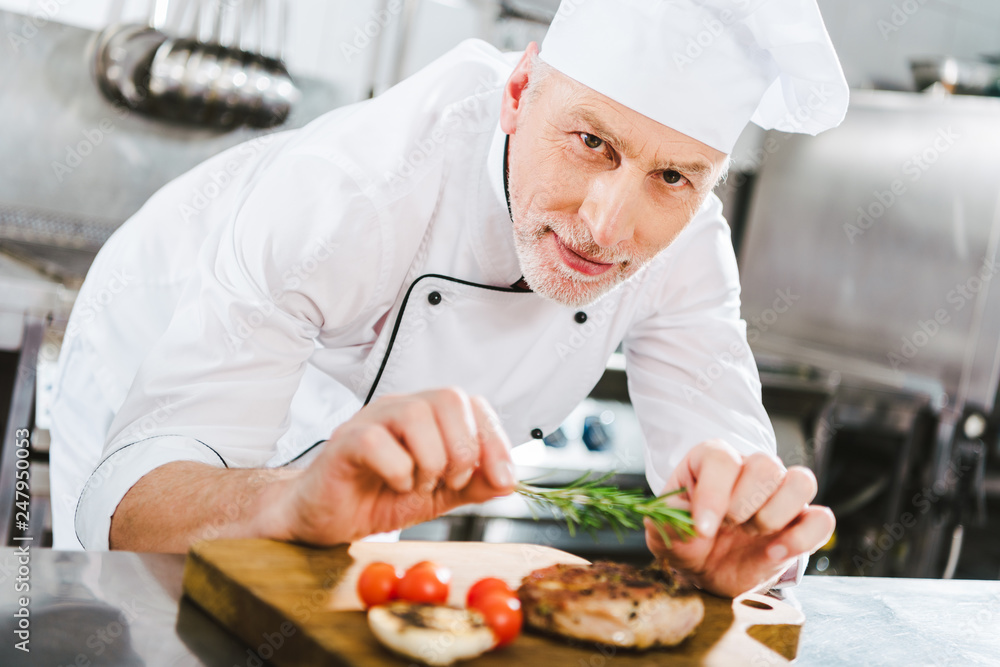 male chef in uniform decorating meat steak with rosemary and looking at camera in restaurant kitchen