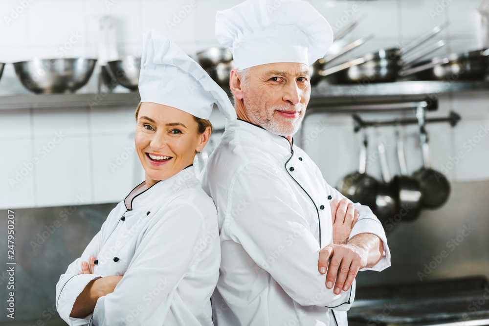 female and male chefs in uniform with arms crossed looking at camera at restaurant kitchen