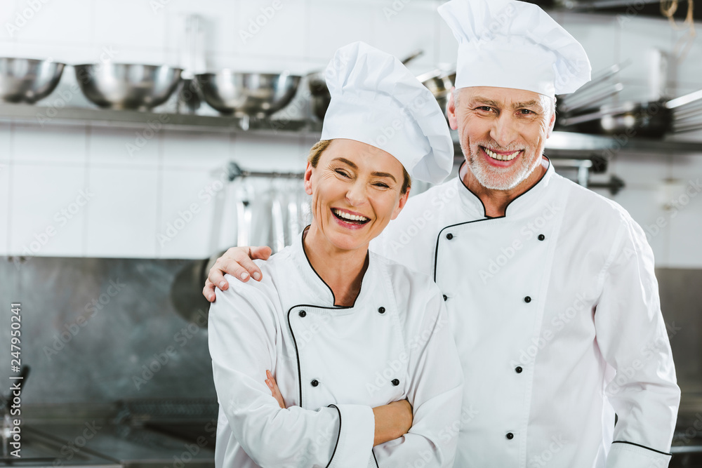 female and male chefs in uniform looking at camera and laughing at restaurant kitchen