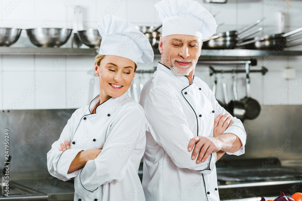 smiling chefs in uniform with arms crossed at restaurant kitchen