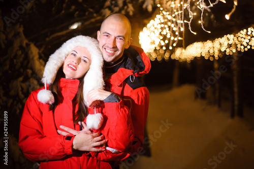 Portrait of happy cute married couple hugging each other while walking at christmas eve outdoors, many lights on the background.