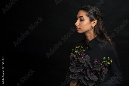 Sad woman wearing a black dress holding black orchid against black background. Copy Space