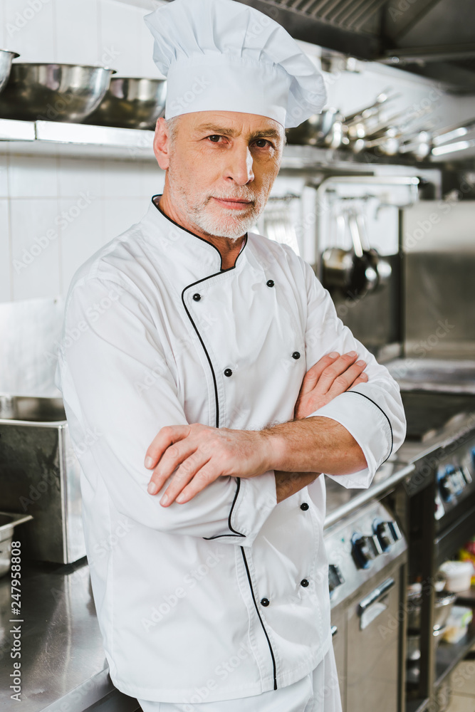 handsome male chef in uniform with arms crossed looking at camera in restaurant kitchen