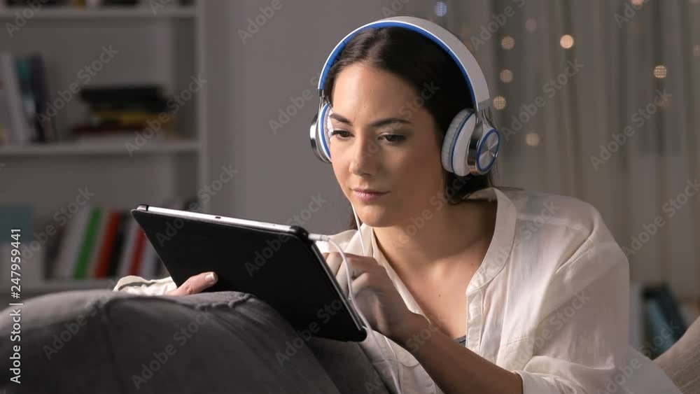 Serious girl with headphones searching media on tablet sitting on a couch in the night at home