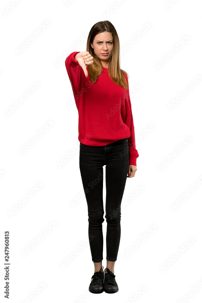 © luismolinero - A full-length shot of a Young woman with red sweater showing thumb dowg with negative expression over isolated white background