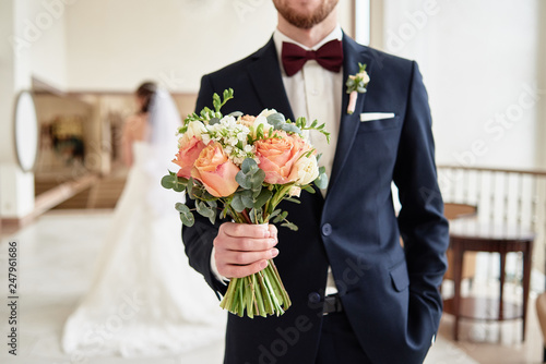 Fotografie Close up of beautiful bridal bouquet of coral roses and greenery in groom's hand with bride in blur on background indoors, copy space