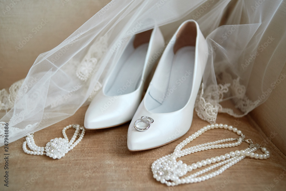 Luxury white shoes on high heels, veil, golden wedding rings and bride jewelry (pearls necklace and bracelet) on chair, copy space. Wedding morning preparation. Bridal accessories. Marriage concept Luxury white shoes on high heels, veil, golden wedding rings and bride jewelry (pearls necklace and bracelet) on chair, copy space. Wedding morning preparation. Bridal accessories. Marriage concept