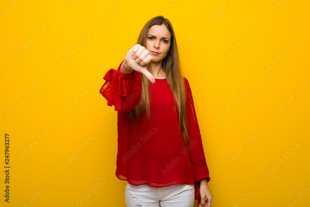 Young girl with red dress over yellow wall showing thumb down sign with negative expression Young girl with red dress over yellow wall showing thumb down sign with negative expression