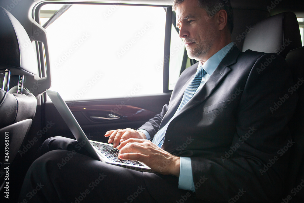 Handsome businessman sitting with laptop on the backseat of the car.