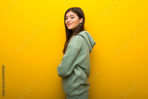 Teenager girl with green sweatshirt on yellow background keeping the arms crossed in lateral position while smiling © luismolinero