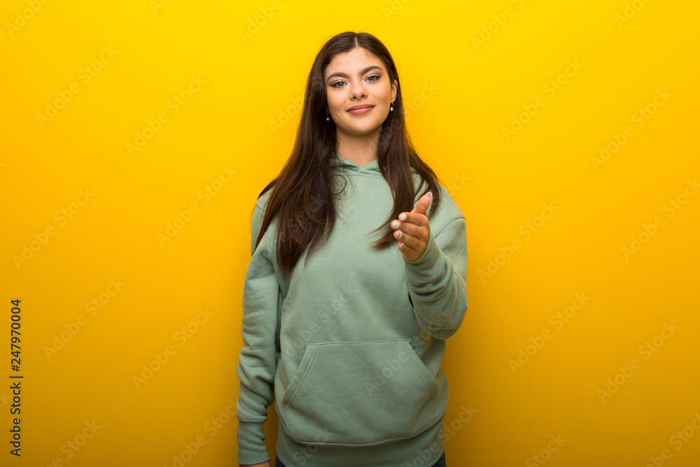 Teenager girl with green sweatshirt on yellow background shaking hands for closing a good deal Teenager girl with green sweatshirt on yellow background shaking hands for closing a good deal