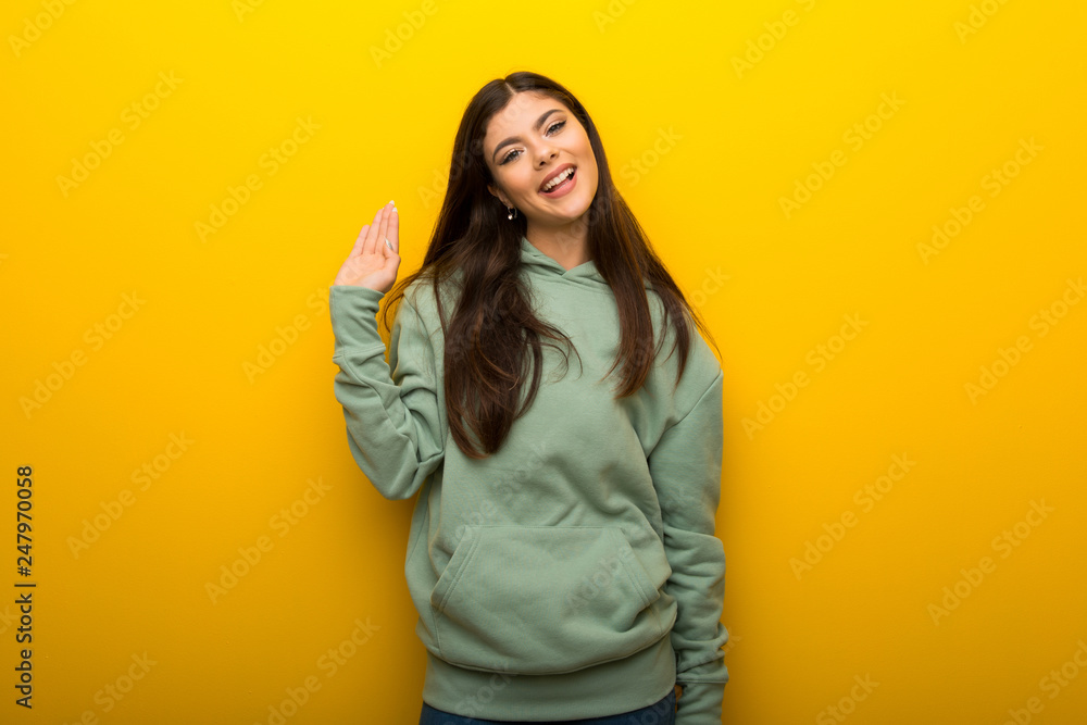 Teenager girl with green sweatshirt on yellow background saluting with hand with happy expression Teenager girl with green sweatshirt on yellow background saluting with hand with happy expression