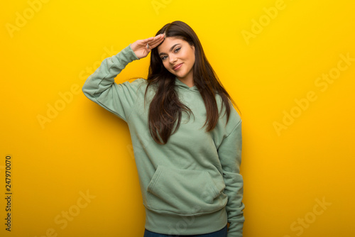 Teenager girl with green sweatshirt on yellow background saluting with hand © luismolinero