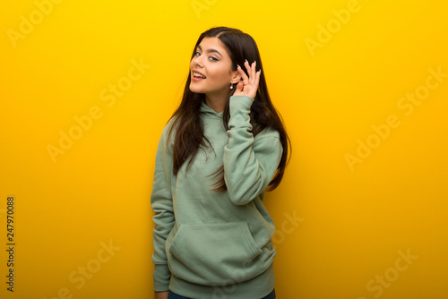 Teenager girl with green sweatshirt on yellow background listening to something by putting hand on the ear © luismolinero