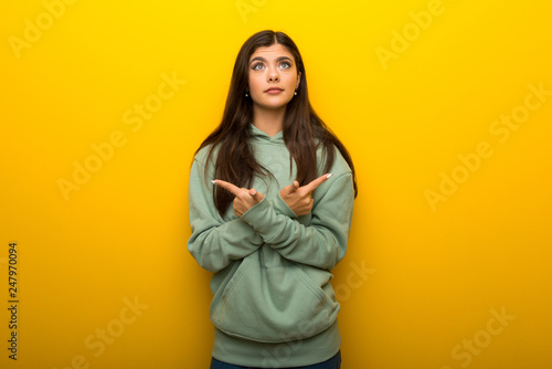 Teenager girl with green sweatshirt on yellow background pointing to the laterals having doubts