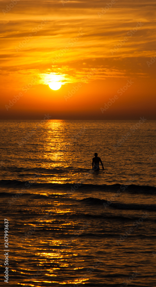 Silhouette of the young man walking in the sea toward the golden sunset with saturated sky and clouds. Beautiful seascape in the evening. Harmony with nature idea. Tranquility and freedom background.