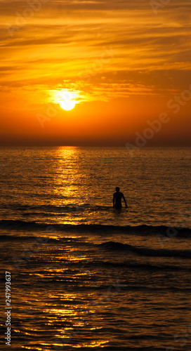 Silhouette of the young man walking in the sea toward the golden sunset with saturated sky and clouds. Beautiful seascape in the evening. Harmony with nature idea. Tranquility and freedom background.