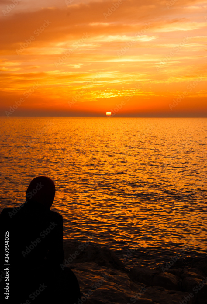 Silhouette of Arab woman in traditional Muslim black hijab or jilbab looking on the beautiful golden sunset in the sea with saturated sky and clouds. Back view. Peaceful serene landscape. Middle East.