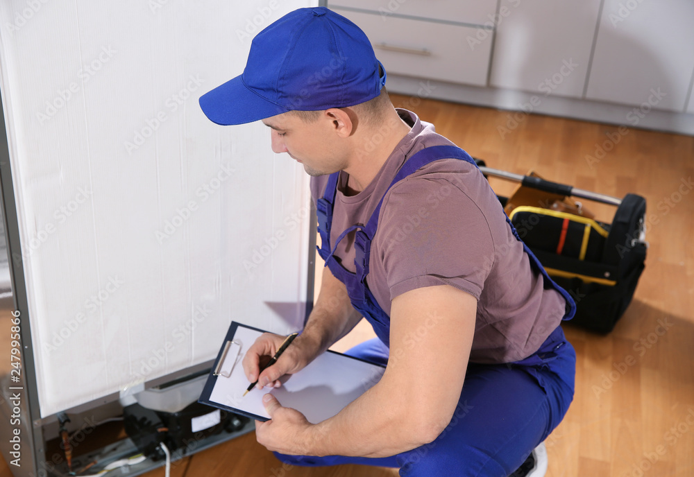 Male technician with clipboard examining refrigerator in kitchen Male technician with clipboard examining refrigerator in kitchen