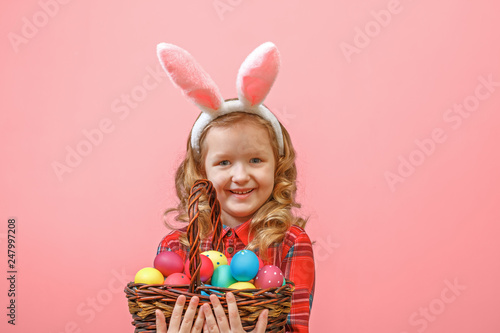 Cute little child girl with bunny ears holding basket of Easter eggs on a colored background.