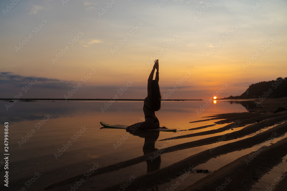 Silhouette and reflection of girl sitting on surfboard at ocean beach on background of beautiful sunset