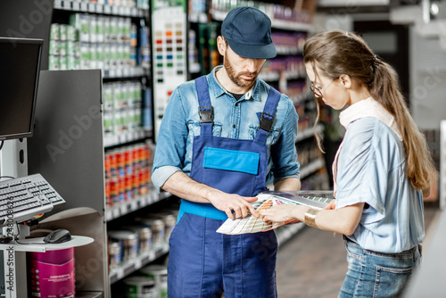 Fototapeta Naklejka Na Ścianę i Meble -  Young woman client choosing paint from color swatches, standing with workman in supermarket with building goods