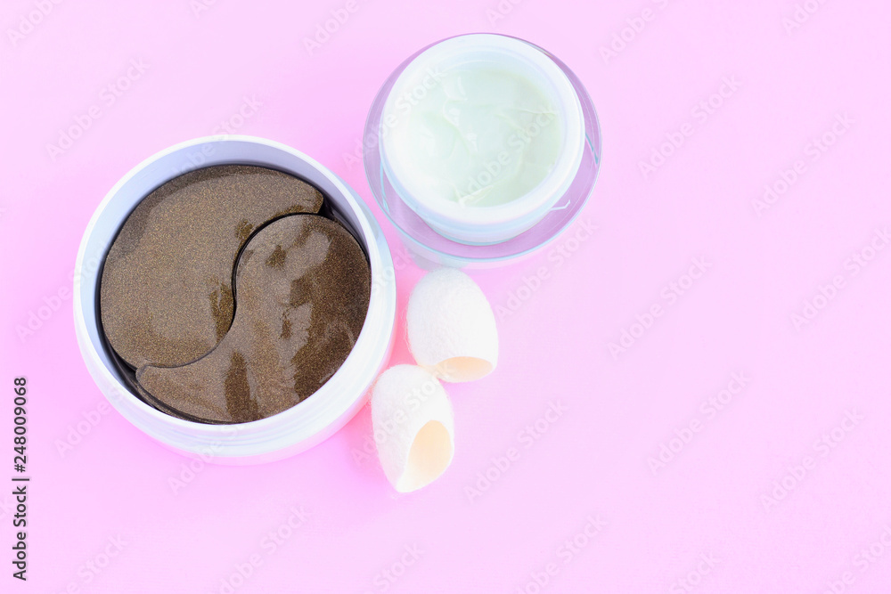 Green face cream and container with black golden anti aging collagen lifting patches under eyes for woman face care and pure silky fiber cocoons with selective focus on pink neutral background Green face cream and container with black golden anti aging collagen lifting patches under eyes for woman face care and pure silky fiber cocoons with selective focus on pink neutral background