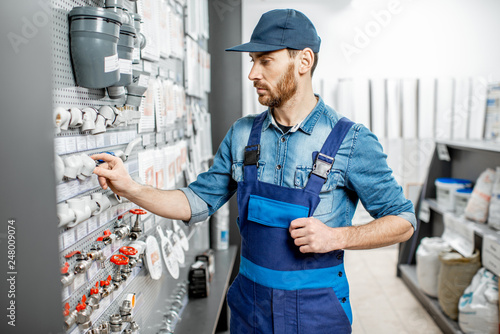 Handsome workman choosing water pipes and pipe joints standing near the showcase in the plumbing shop