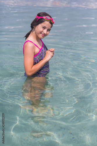 a little girl in a swimsuit and glasses for swimming on her head stands waist-deep in clean sea water and looks at the camera © Nikita