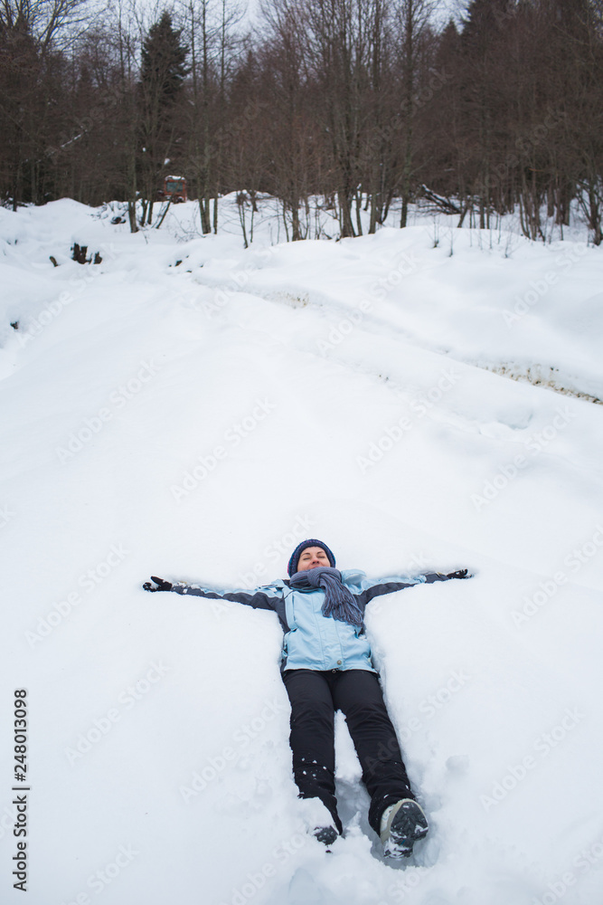 girl in pants, jacket, gloves and scarf lay in the snow in front of the forest macking snow angel