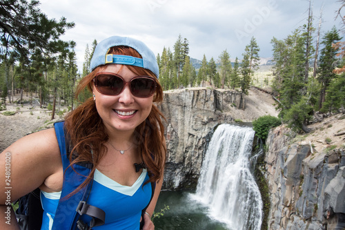 Redhead female hiker wearing a ballcap poses at Rainbow Falls in California along the John Muir Trail photo