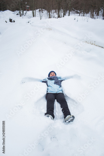 girl in pants, jacket, gloves and scarf lay in the snow in front of the forest macking snow angel