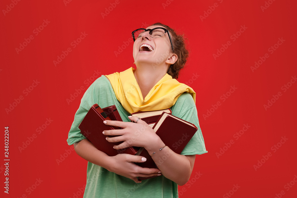 Positive young student with glasses and books in her hands laughs with her head thrown back depicting cheerfulness on a red background. Advertising space Positive young student with glasses and books in her hands laughs with her head thrown back depicting cheerfulness on a red background. Advertising space
