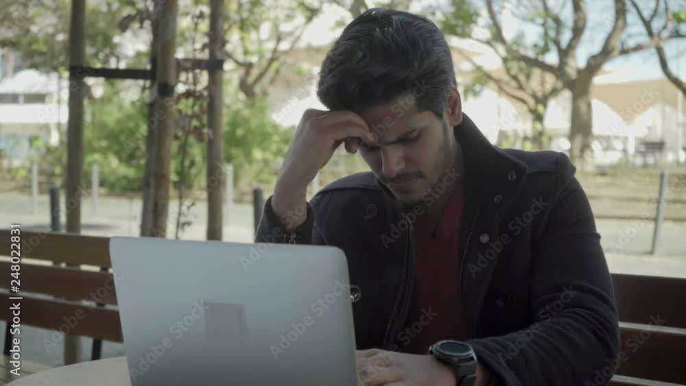 Serious young man thinking and working with laptop. Focused busy freelancer sitting on bench and using laptop computer outdoor. Difficult task concept Serious young man thinking and working with laptop. Focused busy freelancer sitting on bench and using laptop computer outdoor. Difficult task concept