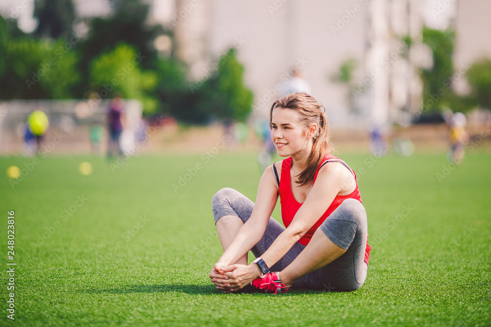 Theme sport and health. Young beautiful Caucasian woman sitting doing warm-up, warming up muscles, stretching green grass. Restoration and rest of girl on stadium in summer on artificial turf Theme sport and health. Young beautiful Caucasian woman sitting doing warm-up, warming up muscles, stretching green grass. Restoration and rest of girl on stadium in summer on artificial turf
