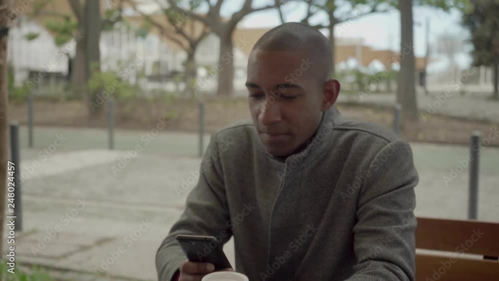 Man holding paper cup and using smartphone on street. Vertical motion of handsome African American man in coat sitting on bench and texting via cell phone. Technology concept Man holding paper cup and using smartphone on street. Vertical motion of handsome African American man in coat sitting on bench and texting via cell phone. Technology concept