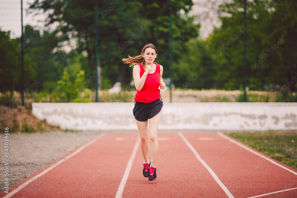 Beautiful young athlete Caucasian woman with big breasts in red T-shirt and short shorts jogging, running in the stadium with red rubber coating