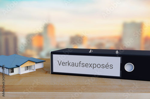 Exposé für Immobilien-Verkauf. Ordner beschriftet mit dem Wort Verkaufsexposés liegt neben einem Haus-Modell auf einem Schreibtisch. Skyline einer Stadt im Hintergrund. photo