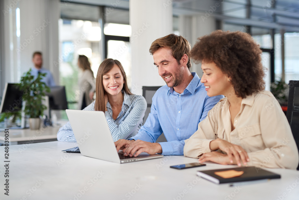 Coworkers sitting at table and solving problem. Man using laptop while two female colleagues looking at laptop and helping him..