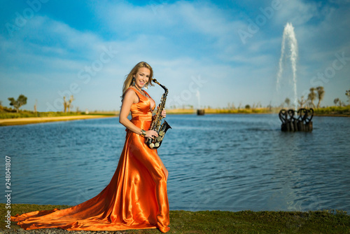 Girl with sax smiles, flirts. Happy blonde woman with big bust plays, performs on goldy saxophone in beautiful dress photo