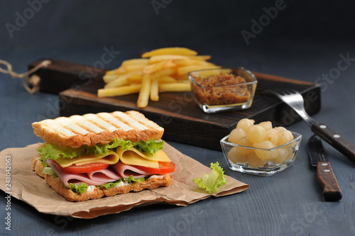 Sandwich with ham and cheese on craft paper. In the background are french fries, pickled onions, mustard and cutlery. Dark background Close-up.