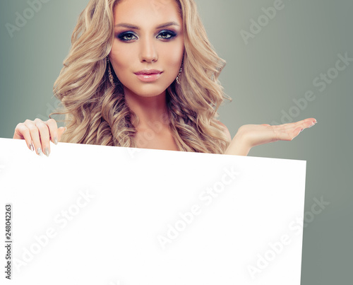 Blonde fashion model girl holding white empty paper board background. Beautiful blonde woman
