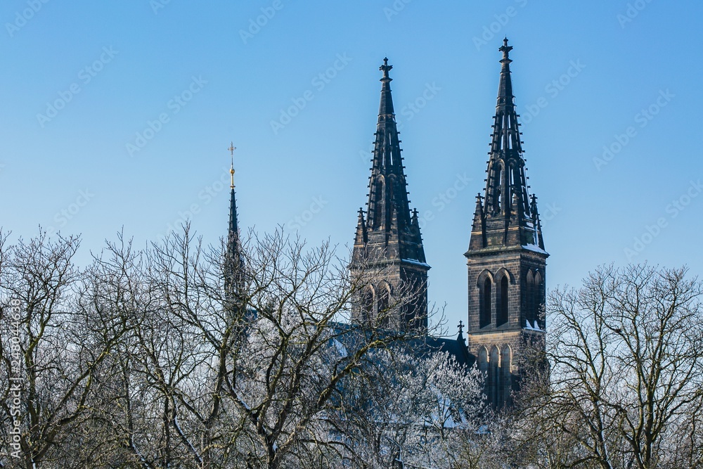 Prague, Czech Republic / Europe - February 5 2019: Towers of Basilica of Sts Peter and Paul at Vysehrad made of stone, spire with golden cross, blue sky, sunny winter day, trees covered with snow