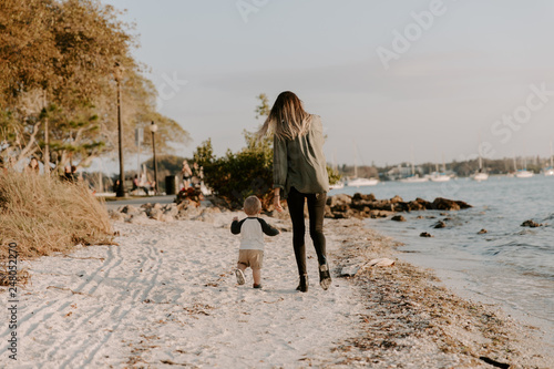 Beautiful Young Mother and Cute Little Boy Son Walking and Enjoying the Nice Outdoor Weather on the Sandy Beach next to the Ocean Bay Water in Summer Outside