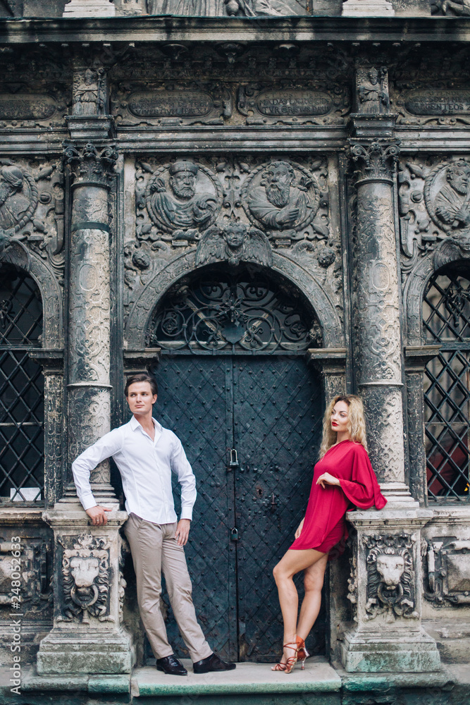beautiful happy couple in love posing at old building in sunny street. stylish hipster groom and blonde bride in red dress gently hugging, professional dancers. romantic moments in summer